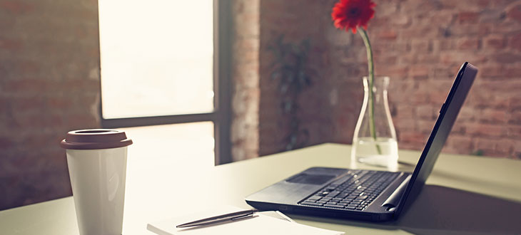 desk with pen and paper, open laptop, coffee cup, and carnation in vase