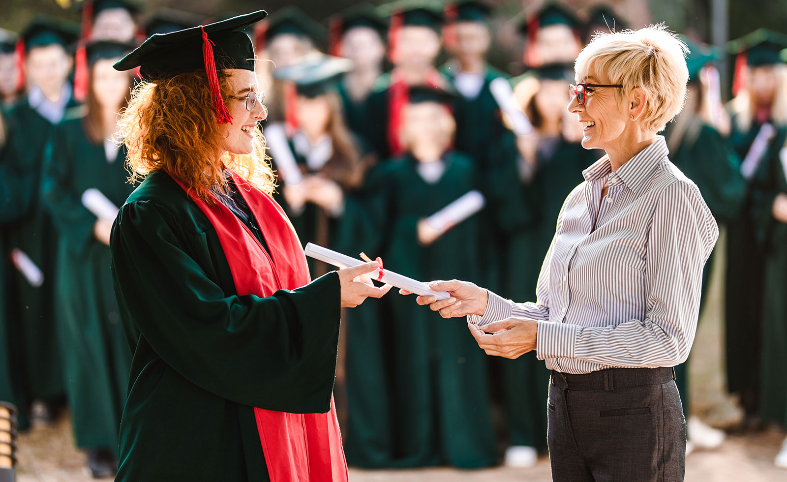 Young woman wearing a graduation cap and gown stands in line with her graduating peers holding her diploma.