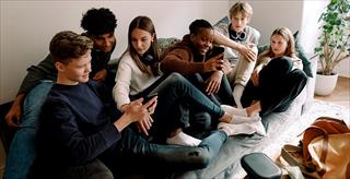 Six teenagers of various races and ethnicities relaxing on a couch while watching videos on their smart phones.