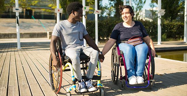 Young, African American man and young, white woman both seated in wheelchairs holding hands.