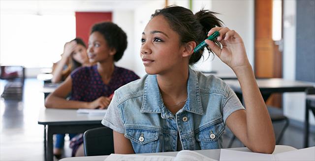 Young female student sitting at desk, holding a green pen to her ear.