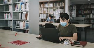 Student sitting in library using a laptop and wearing a mask