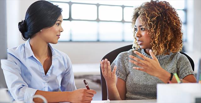 Two professional women sitting at a desk and exchanging spoken information