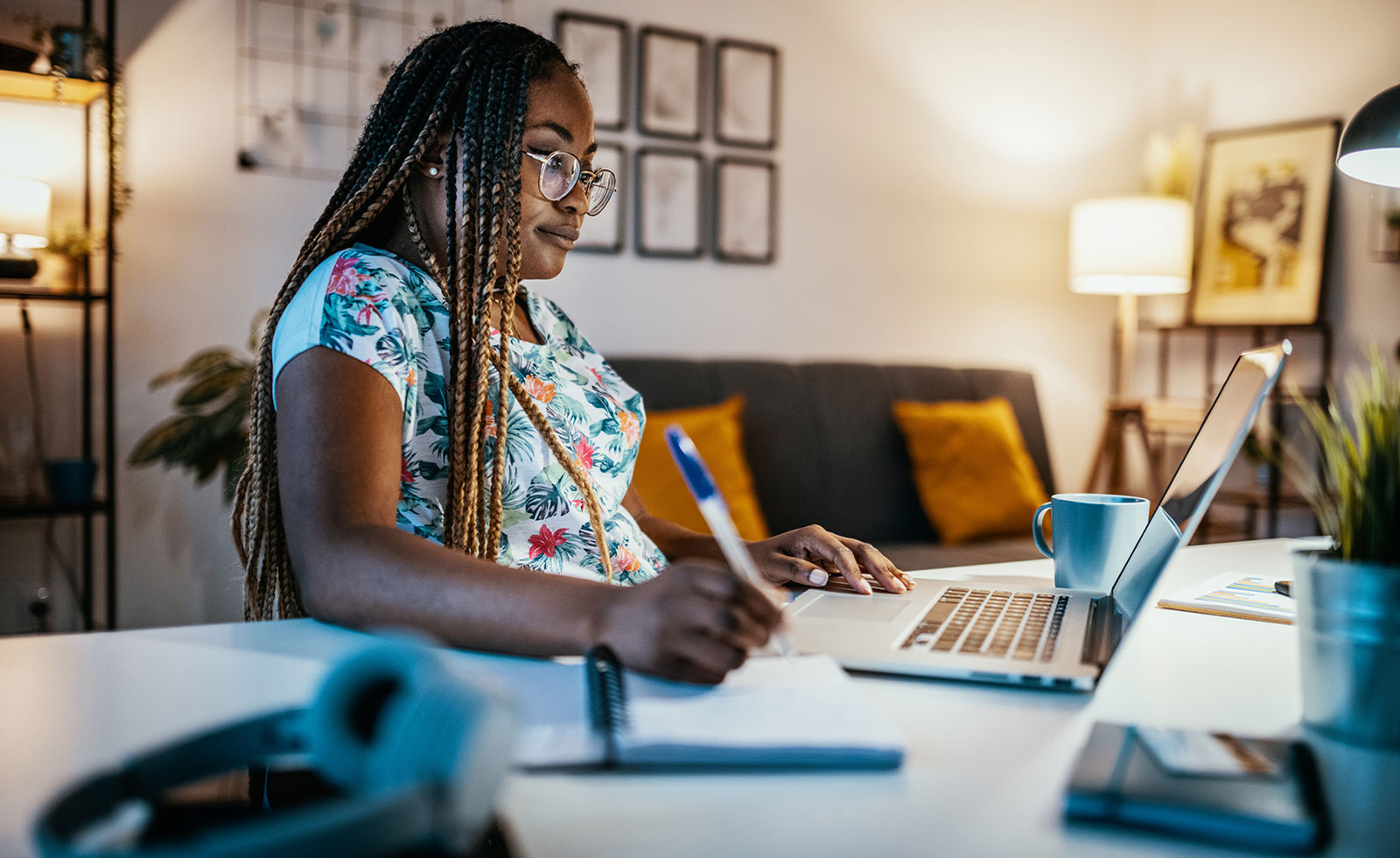 Black female student writing notes while looking at laptop.