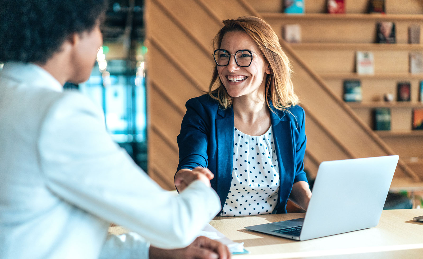 smiling woman sitting in front of a laptop shaking hands with another seated woman